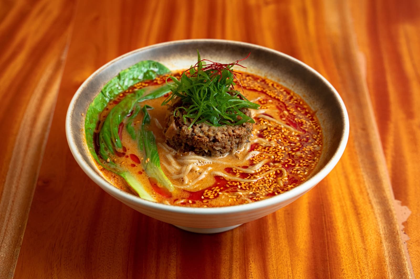 A bowl of ramen on the wooden counter: minced meat and shredded spring onion over the noodles, a leaf of greens at the side, sesame across a red broth