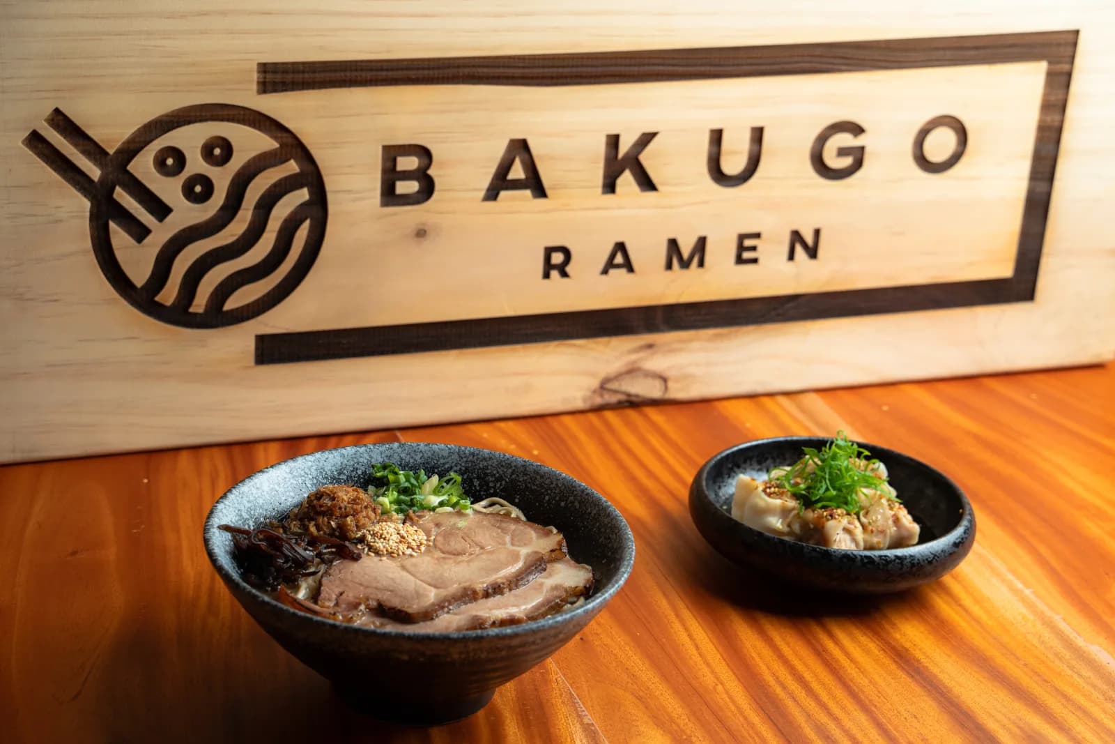 The carved Bakugo Ramen sign behind two bowls on the wooden counter